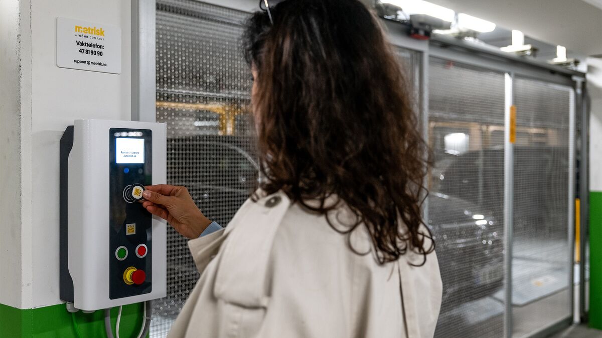 En person betjener kontrollpanelet til et halvautomatisk parkeringssystem for å hente et parkert kjøretøy. I bakgrunnen er de flyttbare parkeringsplassene og et kjøretøy inne i parkeringsanlegget synlige.